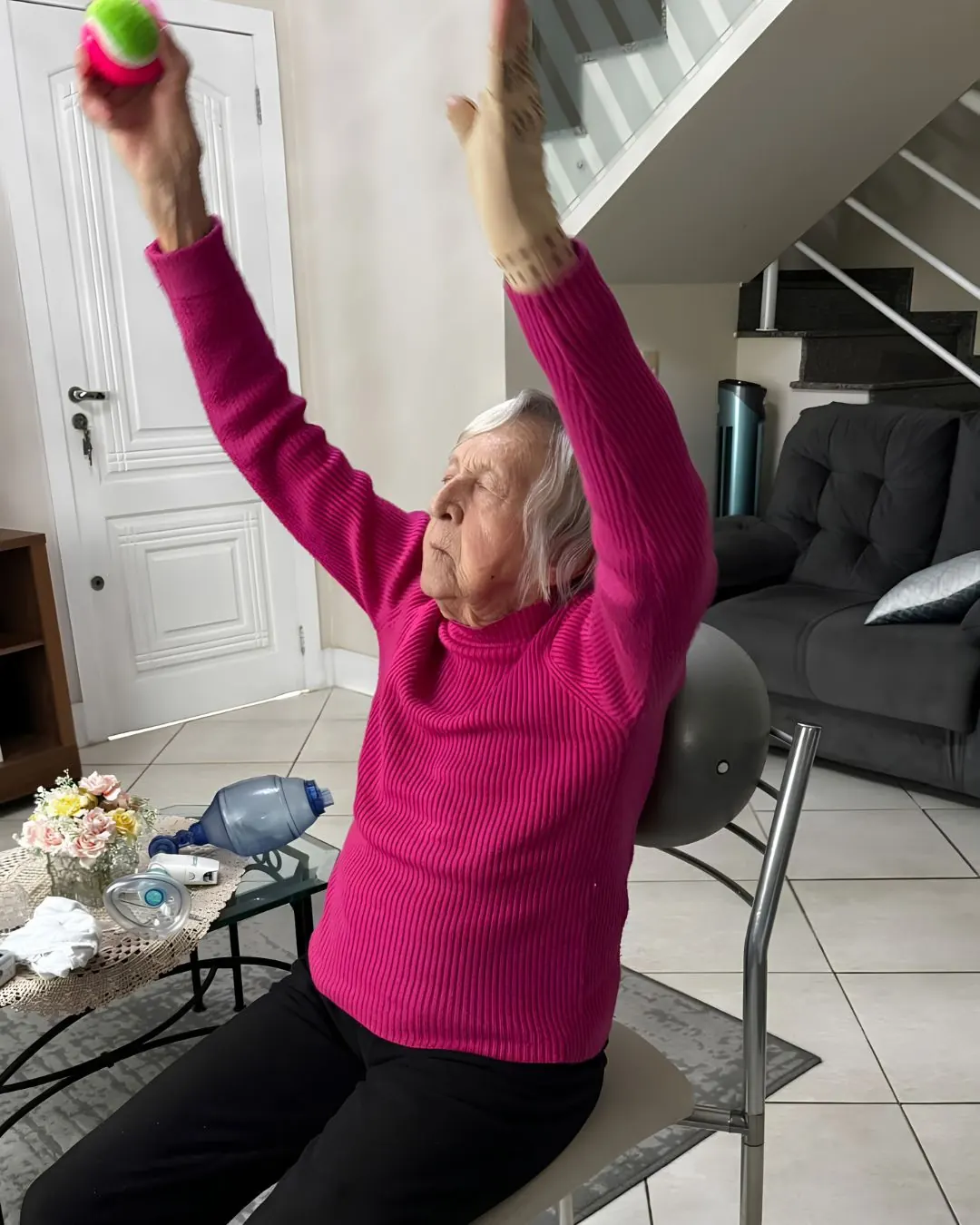 Paciente idosa realizando exercício de fortalecimento com peso, sentada em casa.