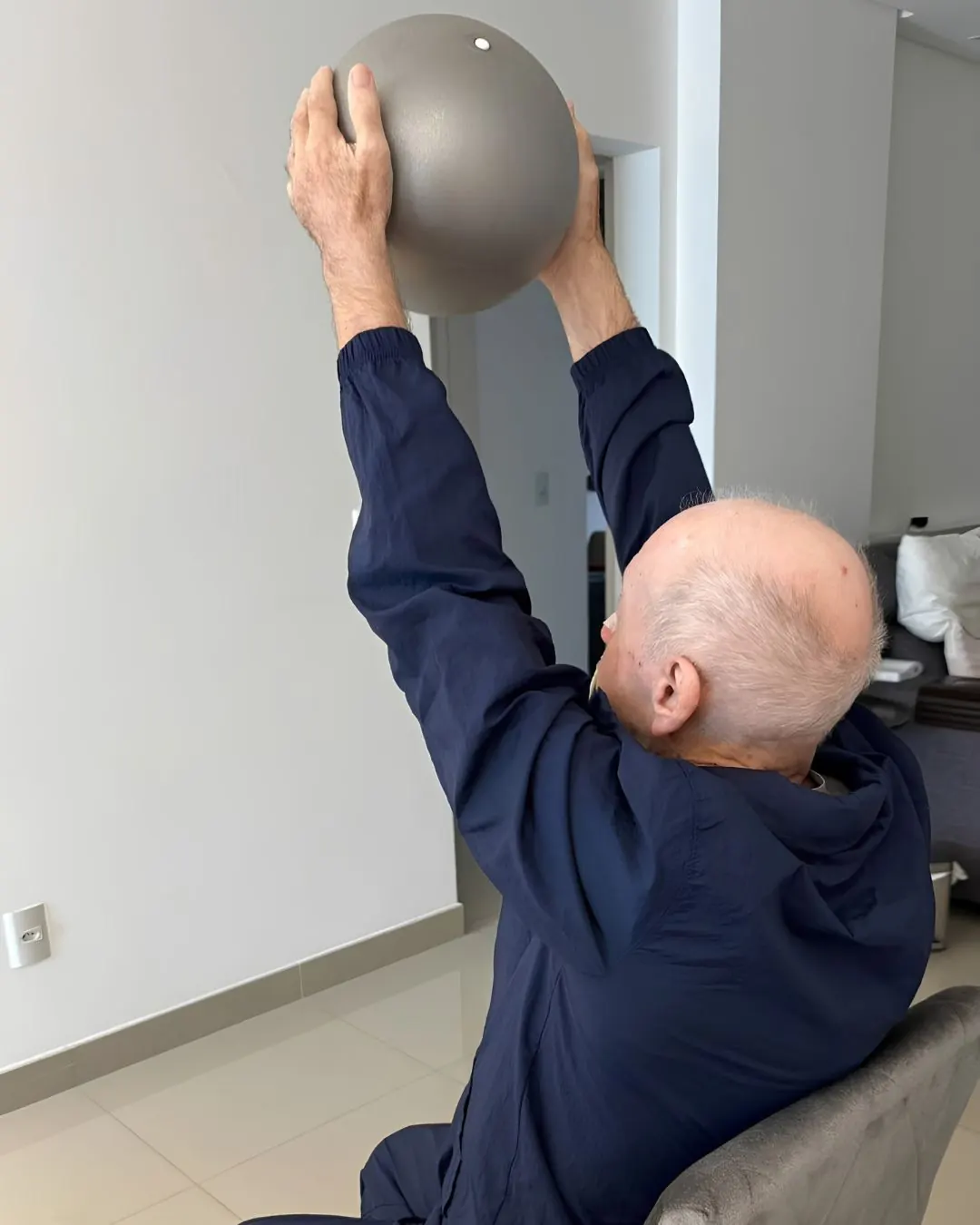 Paciente idoso realizando exercício de fortalecimento com bola, em atendimento domiciliar.
